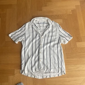 Vit randig skjorta från Zara, M - Snygg vit skjorta från Zara med svarta vertikala ränder. Skjortan har korta ärmar, klassisk krage och knäppning framtill. Tillverkad i ett luftigt linnematerial som passar perfekt till varmare dagar. Enkel och stilren design som är lätt att matcha.