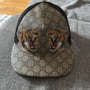 Gucci keps med tigertryck och GG-mönster - Snygg Gucci keps i beige med klassiskt GG-mönster och två stora tigertryck framtill. Baksidan är i svart mesh med justerbart band i grönt och rött. Kepsen är tillverkad i bomull och polyester, perfekt för dig som vill sticka ut med lyxig streetstyle.