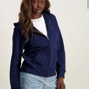 Mörkblå hoodiejacka från Polo Ralph Lauren - Mörkblå hoodiejacka från Polo Ralph Lauren med dragkedja framtill, huva och kängurufickor. Klassisk röd logga broderad på bröstet. Skön bomullskänsla och långärmad modell, perfekt till jeans eller joggers.