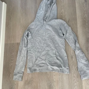 Grå hoodie från Svea med broderad logga - Säljer en grå hoodie från Svea med broderad logga framtill. Tröjan har huva med snörning och en stor magficka. Perfekt för chill dagar och enkel att matcha med jeans eller mjukisbyxor.