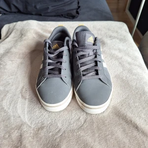 Grå Adidas sneakers med vita ränder - Snygga grå sneakers från Adidas med klassiska vita ränder på sidorna och gulddetaljer på häl och plös. Skorna har snörning, platt sula och rund tå. Perfekta för dig som gillar en clean och sportig stil. Knappt använda 