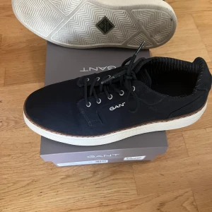 Marinblå sneakers från Gant - Stilrena marinblå sneakers från Gant med vit sula och snygga detaljer i brunt längs kanten. Skorna har snörning med metallöljetter och ovandel i textil. Perfekta för dig som gillar en klassisk men modern look. Endast använda ett fåtal gånger och nypris-1400kr. Skriv till mig privat vid frågor eller funderingar! 