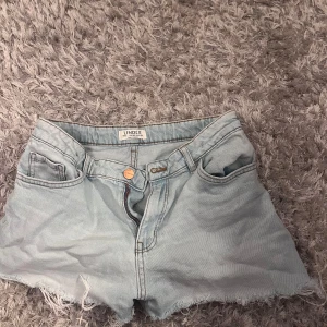 Ljusblå jeansshorts från Lindex - Snygga ljusblå jeansshorts från Lindex med råa kanter nertill och klassisk femficksdesign. Shortsen har bälteshällor, knapp och dragkedja framtill. Perfekta för varma dagar och enkel att matcha med allt.