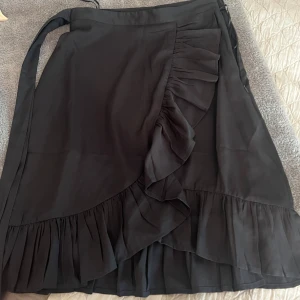 Svart omlottkjol med volang - Snygg svart omlottkjol med bred volangkant och knytband i midjan. Kjolen har en flowy känsla och volanger längs nederkanten och framtill, vilket ger en trendig och feminin look. Perfekt att styla med boots eller sneakers.