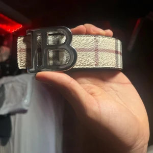  Burberry bälte - Snyggt bälte från Burberry med klassiskt rutigt mönster i beige, rött och grått. Bältet har ett stort metallspänne formad som ett B och är gjort i skinn. Perfekt accessoar för att lyfta din outfit.