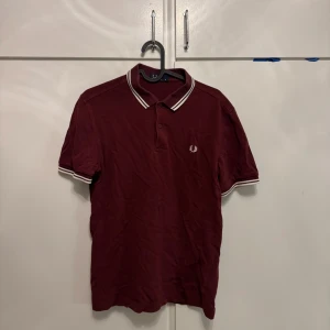 Vinröd Fred Perry pikétröja M - Snygg vinröd pikétröja från Fred Perry med klassisk krage och vita ränder på krage och ärmslut. Ikonisk logga broderad på bröstet. 