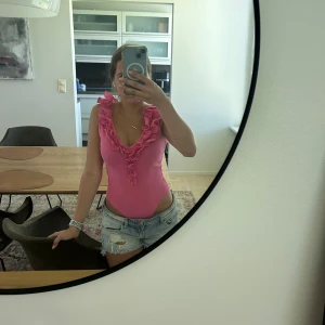 Rosa volangbody - Supersöt rosa ribbad topp från MB-21 Collection med volangdetaljer längs djup v-ringning och axlar. Ärmlös och figurnära passform, perfekt för att sticka ut med en färgglad vibe. Materialet är stretchigt och bekvämt.
