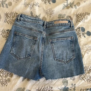 Blå jeansshorts från LAB Industries - Snygga blå jeansshorts från LAB Industries med råa, fransiga kanter och klassisk femficksdesign. Shortsen har normal passform, bälteshällor och dragkedja med knapp. Perfekta för varma dagar och en avslappnad look.