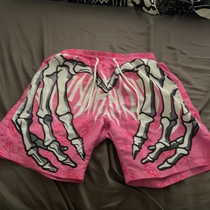 Rosa shorts med skelett-tryck - Unika rosa shorts med paisleymönster och stort vitt och svart skelett-tryck på framsidan. De har elastisk midja med snörning och är tillverkade i ett lätt, syntetiskt material. Perfekta för dig som vill sticka ut med en cool och edgy stil.