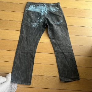 Svarta jeans med blått mönster BX - Unika svarta jeans från BX med ljusblå broderade detaljer framtill och bak, samt tryck över bakfickorna. Jeansen har rak passform och klassisk femficksdesign. Materialet är denim och färgen är tvättad svart med coola blå inslag.