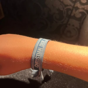 Blått Christian Dior J'Adior armband - Supersnyggt blått armband från Christian Dior med texten 'CHRISTIAN DIOR J'ADIOR' broderad i vitt. Armbandet är gjort i tyg med justerbar knytning och coola tofsar i ändarna. Perfekt accessoar för att lyfta din outfit med en lyxig touch.