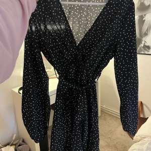 Svart prickig omlottklänning -  svart klänning med vita prickar och knytband i midjan. Klänningen har långa ärmar och v-ringad hals, perfekt för dig som gillar en klassisk men trendig stil.