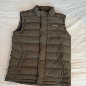 Olivgrön dunväst från GANT - Snygg olivgrön dunväst från GANT med quiltad design och hög krage. Västen har två snedställda fickor framtill och stängs med dragkedja. Insidan är mörkblå och materialet är mjukt och lätt, perfekt för lager-på-lager-stil.