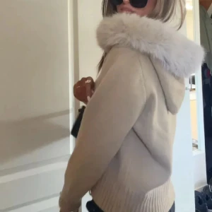 Beige cardigan jacka med fuskpäls från Alessa - Supermysig beige cardigan jacka från Alessa med fluffig fuskpäls runt luvan. Jackan har dragkedja framtill, ribbade muddar och är långärmad. Perfekt för kalla dagar när du vill vara både varm och snygg.