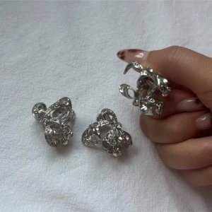 Silvriga rosett hårklämma med strass - Tre hårklämmor i form av glittriga rosetter i silverfärg med små strasstenar och hängande detaljer. De har en snygg, öppen design och gnistrar fint i ljuset. Perfekta för dig som vill ha något extra till din look.
