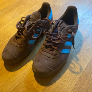Adidas Gazelle bruna och blå sneakers - Säljer ett par trendiga Adidas Gazelle sneakers i brunt mocka med klassiska blå ränder och blå häl med vit logga. Endast testade!!
