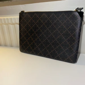 Kuvertväska By Malene Birger brun - Snygg kuvertväska från By Malene Birger i brunt och svart med klassiskt rutmönster. Väskan är i skinnliknande material och har guldfärgad dragkedja. Perfekt storlek för det viktigaste och stilren design som lyfter din look. Helt ny utan band