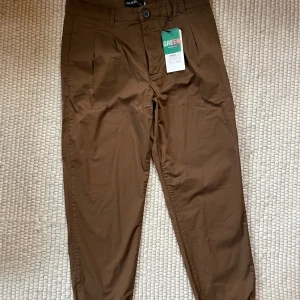 Bruna kostymbyxor från Ljung - Snygga bruna kostymbyxor från Ljung i 100% polyester. Byxorna har klassisk passform med pressveck och stängs med knapp och dragkedja. Perfekta för en clean och stilren look.