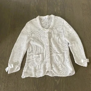 Vit stickad kofta med rosetter - Supersöt vit stickad kofta  med långa ärmar och två fickor framtill. Koftan har fina rosettdetaljer vid halsen och vid ärmsluten som ger en gullig touch. Perfekt att slänga över en topp när det är lite kyligt ute. Obs dåliga bilder blev det men den är i nyskick utan fläckar eller nått❤️