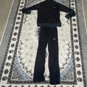 Svart grå tracksuit från Lacoste - Säljer en svart quiltad tracksuit från Lacoste i storlek S. Setet består av en jacka med dragkedja och matchande byxor med snörning i midjan. Båda delarna har den klassiska gröna Lacoste-loggan. Materialet är mjukt och bekvämt. Går också köpa enkilda plagg. .