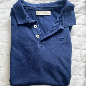 Blå piké från Zara - Klassisk marinblå pikétröja från Zara med krage och tre knappar framtill. Tröjan har korta ärmar och diskret broderad logga på bröstet. Perfekt för en clean och stilren look, passar till både jeans och shorts.