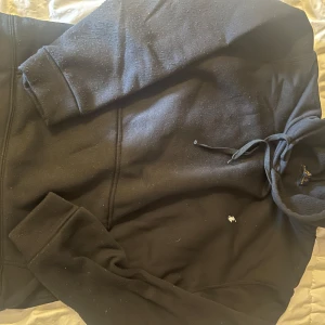 Svart hoodie från Polo Ralph Lauren - Svart hoodie från Polo Ralph Lauren med klassisk vit broderad logga på bröstet. Tröjan har huva med snörning och en stor magficka. Mjuk insida. Perfekt för en chill och stilren look.
