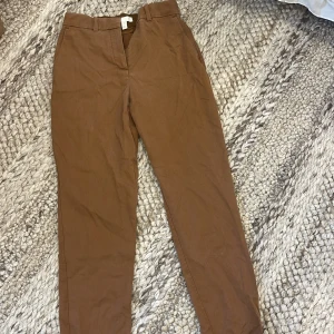 Bruna chinos från H&M, stl 34 - Snygga bruna chinos från H&M i storlek 34. Byxorna har klassisk rak passform, bälteshällor och sidfickor. 