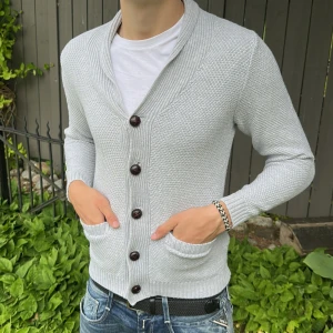 Ljusgrå stickad cardigan  - Säljer en ljusgrå stickad cardigan med stora mörka knappar och två fickor framtill. Cardiganen har lång ärm och en bred ribbad krage som ger en snygg och avslappnad look. Perfekt att slänga på sig över en t-shirt.