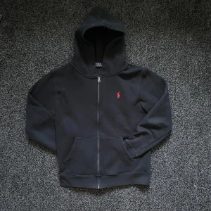 Svart hoodie från Polo Ralph Lauren - Svart hoodie från Polo Ralph Lauren med dragkedja och huva. Klassisk röd logga broderad på bröstet och stora fickor framtill. Tillverkad i mjuk bomull, perfekt för chill dagar. Storlek M. Hör av er vid frågor och funderingar.!köpt begagnad fast ny! Riktigt schyst skick.