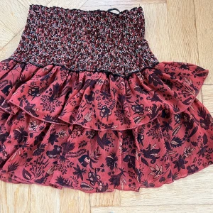 Röd blommig volangkjol med smock - Supersöt kort kjol i röd nyans med svart blommigt mönster och bred smock i midjan. Kjolen har dubbla volanger som ger extra volym och flow. Perfekt för dig som gillar romantisk och bohemisk stil. Materialet känns lätt och luftigt.