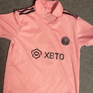 Inter Miami Messi 10 matchtröja rosa - Säljer en rosa Inter Miami fotbollströja från Adidas med Messi och nummer 10 på ryggen. Tröjan har svarta detaljer, klubbmärke på bröstet och sponsortryck framtill. Klassisk kortärmad modell i lätt och ventilerande material, perfekt för fotbollsälskare.