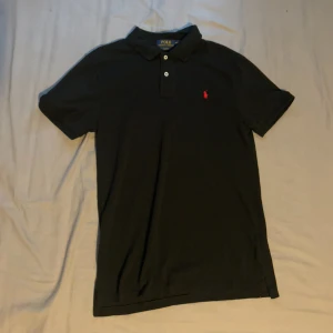 Svart pikétröja Polo Ralph Lauren - Klassisk svart pikétröja från Polo Ralph Lauren med röd broderad logga på bröstet. Slim fit-modell med korta ärmar, krage och två knappar framtill. Tillverkad i mjuk bomull som känns skön mot huden. Perfekt för en clean och stilren look.
