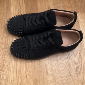 Svarta Louboutin sneakers med nitar - Svarta sneakers från Christian Louboutin i mocka med coola svarta nitar över tån och klassisk röd sula. Kommer med 3 extra nitar OBS saknar en nit.