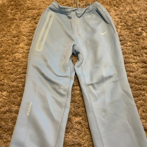 Nike NOCTA ljusblå mjukisbyxor - Snygga ljusblå mjukisbyxor från Nike NOCTA med elastisk midja och dragsko. Byxorna har en dragkedjeficka på sidan, diskret NOCTA-logga och små tryckta detaljer på benen. Perfekta för chill eller streetwear. Mjukt material för maximal komfort.