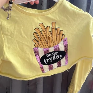 Gul croppad tröja med paljetter - Gul croppad långärmad tröja med ett coolt tryck av pommes och texten 'finally fryday' framtill. Fickan är dekorerad med rosa och vita paljetter för extra bling. Perfekt för dig som gillar statement-plagg och vill sticka ut.