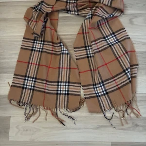 Beige rutig halsduk med fransar - Snygg beige halsduk med klassiskt rutigt mönster i svart, vitt och rött. Halsduken har fransar i ändarna och är perfekt att slänga över axlarna för en cool look. Passar dig som gillar tidlös stil och vill ha något som sticker ut lite extra.