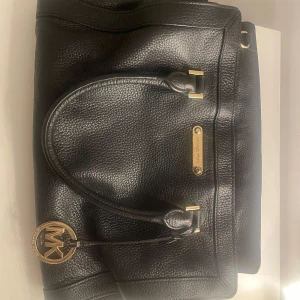 Svart handväska från Michael Kors - Snygg svart handväska från Michael Kors i skinn med guldfärgade metalldetaljer och klassisk MK-logga. 