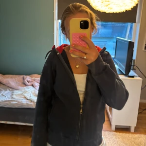 Svart hoodiejacka med röd huva - Svart hoodiejacka med dragkedja och röd insida på huvan. Jackan har långa ärmar, ficka på ärmen och en avslappnad passform. Perfekt för chill dagar eller när du vill ha något bekvämt och snyggt.