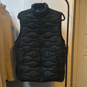 Väst - Snygg svart quiltad väst från Jack & Jones med dragkedja framtill. Perfekt för lager-på-lager under kyligare dagar. Stilren design med hög krage.