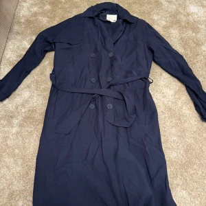 Mörkblå trenchcoat från 157 - Säljer en stilren mörkblå trenchcoat från 157. Jackan har knappar och ett bälte i midjan för en snygg passform. Tillverkad i viskos och polyester, perfekt för vår och höst. Skick: 9/10 Ord. Pris: 239 Storlek: M  Din stil: missa inte chansen! Skriv om du funderar över något. Inte din stil: Kolla in min sida jag har lagt upp klä