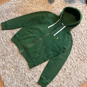 Grön hoodie från Ralph Lauren - Säljer en snygg grön hoodie från Ralph Lauren med dragkedja och huva. Den har en liten gul broderad logga på bröstet och vita snören vid huvan. Perfekt för en avslappnad stil. Har en fläck på baksidan. Fråga bara om mer bilder så löser jag!
