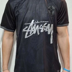 Svart fotbollströja från Stüssy/ Brasilen - Snygg svart fotbollströja från Stüssy med korta ärmar och ett stilrent tryck på framsidan. Perfekt för dig som vill ha en sportig look med en touch av streetwear. Tröjan har en klassisk passform och är tillverkad i ett lätt och bekvämt material.
