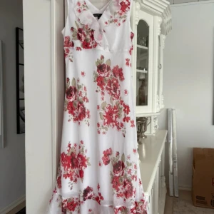 Vit blommig klänning från KappAhl - Säljer en vacker vit klänning från KappAhl med röda blommor och volangdetaljer. Klänningen är ärmlös och har en v-ringad design, perfekt för sommarens varma dagar. Som ny! Storlek M 