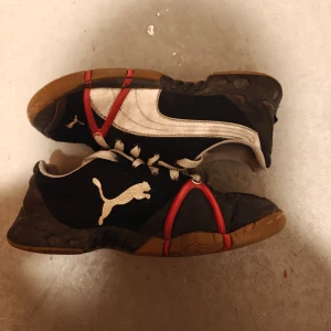 Svarta och vita sneakers från Puma - Säljer ett par svarta och vita sneakers från Puma med röd detalj. Skorna har en klassisk snörning och en robust sula. Perfekta för en sportig look eller vardagsbruk.