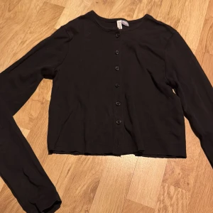 Svart kofta från H&M Divided - Säljer en svart kofta från H&M Divided. Den har långa ärmar och knappar framtill. Perfekt för en stilren look.