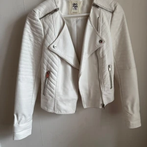Vit skinnjacka från Vero Moda - Snygg vit skinnjacka från Vero Moda med coola dragkedjedetaljer och quiltade partier på axlarna. Jackan har en asymmetrisk dragkedja framtill och två fickor med dragkedjor. Perfekt för att ge din outfit en edgy touch!