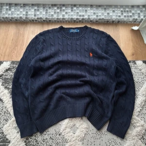 Mörkblå stickad tröja från Ralph Lauren - Säljer en snygg mörkblå stickad tröja från Ralph Lauren med kabelstickat mönster. Tröjan har långa ärmar och en liten broderad logga i rött på bröstet. Perfekt för kyliga dagar! Den har bara varit använd ett fåtal gånger. Pris kan diskuteras och skriv gärna vid intresse!