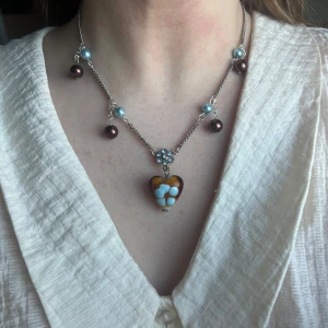 Halsband med hjärthänge och pärlor, vintage - Charmigt halsband med ett hjärthänge i brunt och blått. Kedjan är dekorerad med bruna och ljusblå pärlor som ger en lekfull touch. Perfekt för att lägga till en unik detalj till din outfit. Handgjort och ihopsatt av vintage smycken! 