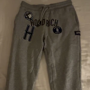 Grå mjukisbyxor från Hoodrich - Säljer min killes gråa mjukisbyxor från Hoodrich med tryck och snörning i midjan. Byxorna har även en diskret ficka med dragkedja vid låret. Perfekta för en bekväm stil. Endast använda ett fåtal gånger och är fria från defekter🥰