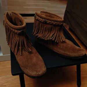 Boots mocka,bruna från Minnetonka - Säljer ett par snygga bruna mocka boots från Minnetonka. De har fransdetaljer och en dragkedja på sidan för enkel på- och avtagning. Perfekta för en bohemisk stil.